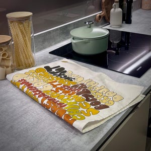 Könnte beinhalten: Eine Küchenszene mit einem weißen Geschirrtuch mit Retro-Design in Braun-, Gelb- und Orangetönen. Das Tuch liegt auf einer Arbeitsplatte neben einem grünen Topf, Glasbehältern mit Pasta und einem schwarzen Kochfeld.
