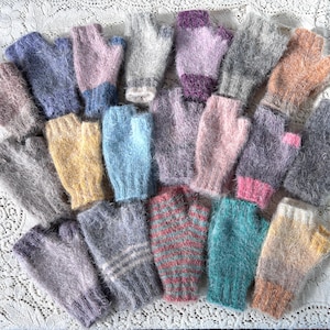 May include: A collection of fingerless gloves in various colors, including blue, pink, yellow, and gray. The gloves are made of a soft, fuzzy material and are arranged on a white lace surface. The gloves are a mix of solid colors and striped patterns.