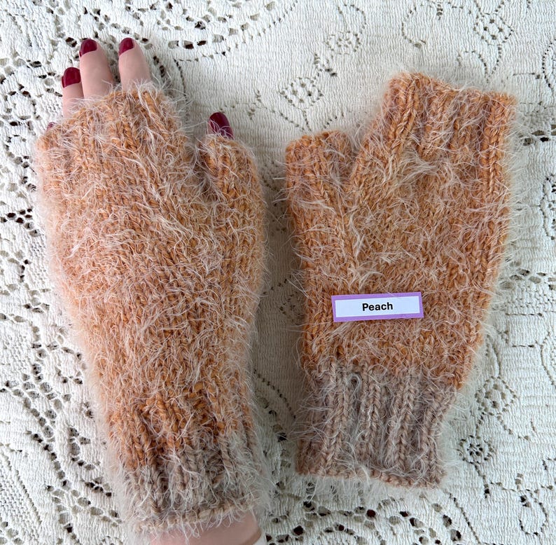 May include: A pair of peach-colored fingerless gloves. The gloves are made of a fuzzy, textured yarn with a ribbed cuff. A small label that says "Peach" is attached to one of the gloves.