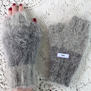 May include: A pair of fingerless gloves in shades of gray, with a fluffy, textured appearance. The gloves are knitted and feature a label that reads "Ash". The overall style is cozy and warm.