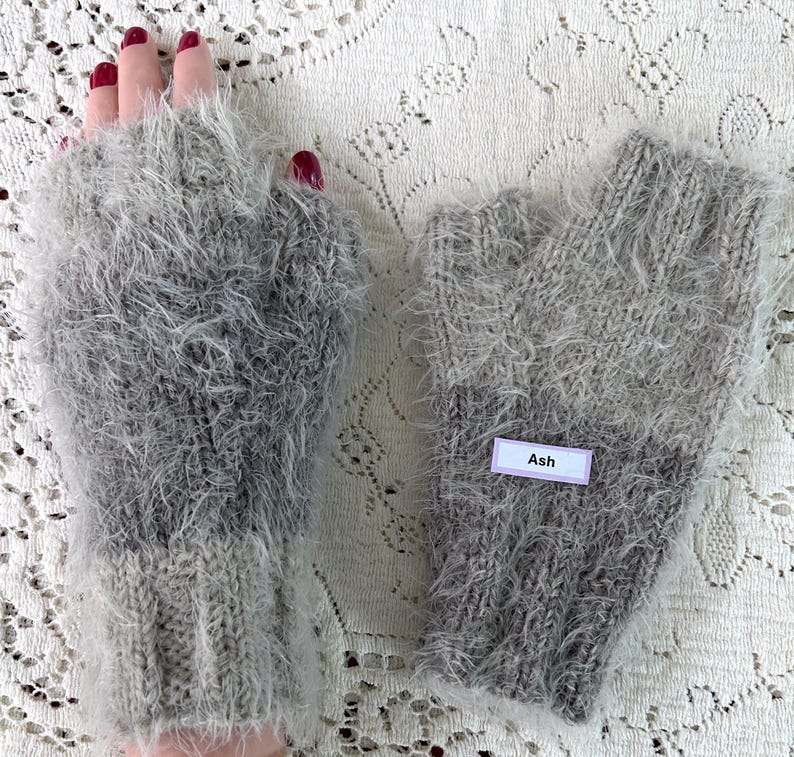 May include: A pair of fingerless gloves in shades of gray, with a fluffy, textured appearance. The gloves are knitted and feature a label that reads "Ash". The overall style is cozy and warm.