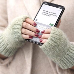 May include: A person holding a smartphone with a text message that reads, "Heading out for lunch, meet you at to the cafe cafe in 15ms!" The person is wearing light green fingerless gloves.