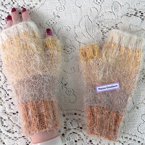 May include: A pair of hand-knitted fingerless gloves with a gradient color scheme. The gloves transition from a light cream color at the top to a soft brown at the wrist, with a fuzzy texture. The label reads "Maize Gradient".