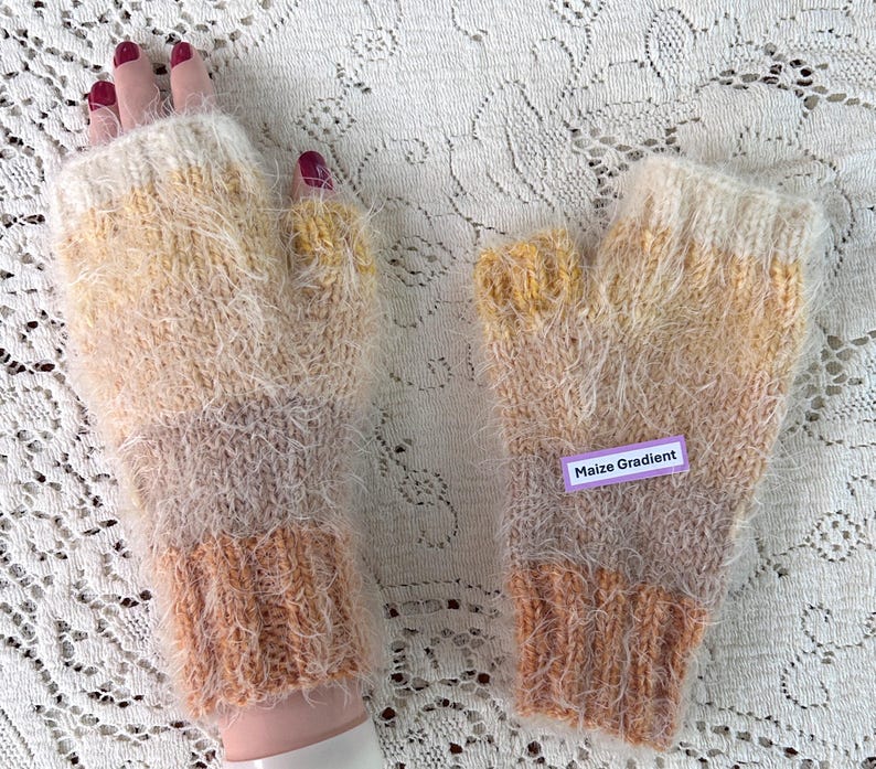 May include: A pair of hand-knitted fingerless gloves with a gradient color scheme. The gloves transition from a light cream color at the top to a soft brown at the wrist, with a fuzzy texture. The label reads "Maize Gradient".
