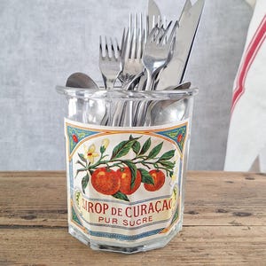 Puede incluir: Un tarro de cristal transparente lleno de cubiertos, incluyendo tenedores, cuchillos y cucharas. El tarro tiene una etiqueta decorativa con tomates rojos y el texto "SIROP DE CURAÇAO PUR SUCRE". El tarro está sobre una superficie de madera.