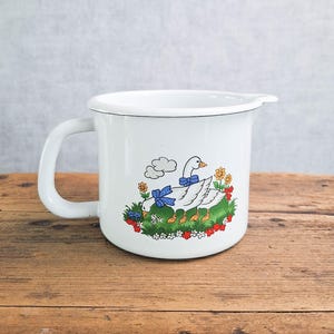 Könnte beinhalten: Ein weißer Emaille-Becher mit einer grafischen Illustration von zwei weißen Gänsen mit blauen Schleifen, die auf einer grünen Wiese mit bunten Blumen stehen. Der Becher hat einen geschwungenen Henkel und einen kleinen Ausguss am oberen Rand.