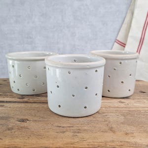 May include: Three off-white ceramic containers with small, evenly spaced holes. The containers are cylindrical with a slightly flared rim. They sit on a weathered wooden surface, with a neutral background and a glimpse of a striped cloth.