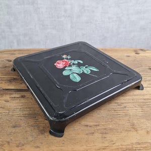 May include: A square black metal trivet featuring a central floral illustration of a single red rose with green leaves and stems. The trivet has a raised border, a smooth finish, and four short, sturdy feet at each corner for elevation.