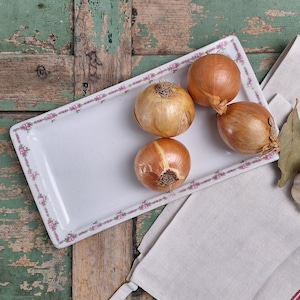 Può includere: Quattro cipolle marroni su un piatto da portata rettangolare bianco con bordo floreale. Un asciugamano di lino con strisce rosse e uno spicchio d'aglio con foglie di alloro accanto al piatto. Lo sfondo è una superficie di legno verniciata di verde.