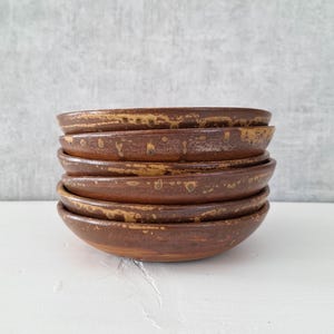 May include: A stack of five circular ceramic bowls with a dark brown base color and a mottled, textured golden-brown glaze along the rims. The bowls are uniform in shape and size, featuring a smooth interior surface and a rustic, earthy appearance.