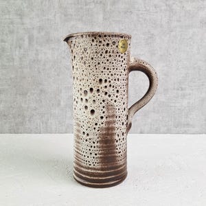 Könnte beinhalten: Hoher Keramikkrug mit gesprenkelter, cremefarbener und brauner Glasur. Der Krug hat einen Henkel und einen Ausguss zum Ausgießen. Das Design weist eine strukturierte Oberfläche mit kleinen, unregelmäßigen Löchern auf. Ein kleiner, goldfarbener Aufkleber ist angebracht.