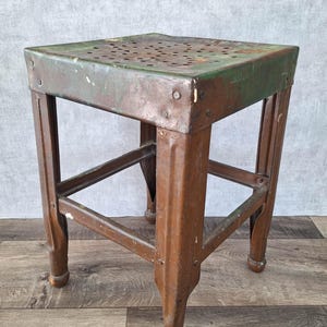 Könnte beinhalten: Ein quadratischer Metallhocker mit einer verwitterten grünen und braunen Patina. Die Sitzfläche hat eine perforierte Oberfläche mit zahlreichen kleinen kreisförmigen Löchern. Der Hocker steht auf vier stabilen, geraden Beinen mit Querstreben.