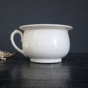 Peut inclure: Un pot de chambre en céramique blanche avec une seule poignée. Le pot est posé sur une surface en bois noir.