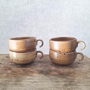 May include: Four ceramic mugs with a speckled tan and brown glaze, arranged in two stacks of two. Each mug features a rounded body, a short handle, and a smooth rim. The glaze displays variations in color density and dark brown speckles.