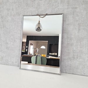 Peut inclure: Un miroir mural rectangulaire doté d'un cadre fin en métal argenté poli. Le miroir est suspendu par une courte chaîne décorative en métal fixée au centre supérieur du cadre, permettant une installation simple et épurée sur un mur.