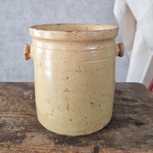 May include: A cylindrical stoneware crock with a light yellow glaze. The crock has two small, rounded handles and subtle horizontal bands. It rests on a weathered wooden surface, suggesting age and rustic charm. The crock's height is approximately 20 cm.