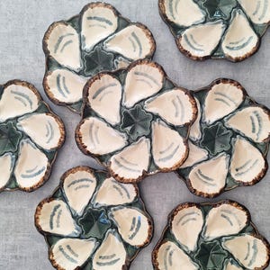 May include: A collection of six ceramic oyster plates, each featuring a circular design with six individual shell-shaped indentations. The plates have a cream-coloured interior with blue accents and a dark green centre, finished with brown edges.