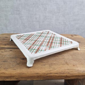 May include: A square metal trivet featuring a white frame and four short legs. The center surface displays a decorative plaid pattern composed of intersecting thin brown and green lines set against a white background, creating a grid-like design.