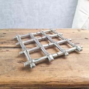 May include: A silver metal trivet with a grid design, resting on a weathered wooden surface. The trivet is made of interconnected bars with decorative joints. The background includes a neutral wall and a white cloth.