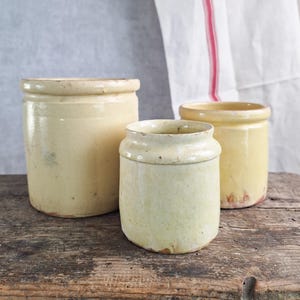 Peut inclure: Trois pots en grès cylindriques antiques de différentes tailles et nuances de jaune et de crème sont posés sur une surface en bois vieilli. Un tissu blanc avec une rayure rouge est suspendu en arrière-plan, ajoutant une touche rustique.