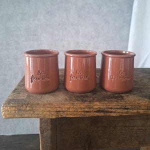 Può includere: Tre vasetti in ceramica color terracotta con la scritta in rilievo "La Fermière". I vasetti sono cilindrici con un bordo leggermente svasato. Sono disposti su una superficie di legno rustico su uno sfondo neutro.