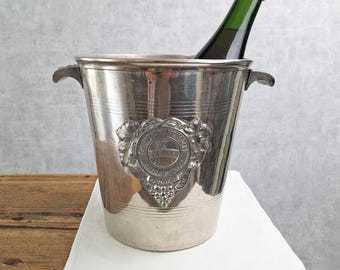 May include: A silver-toned metal champagne bucket featuring two side handles and a central embossed crest with grape vine detailing. A dark green glass bottle with a cork stopper is positioned inside the bucket, which has horizontal lines.