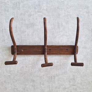 May include: A dark brown wooden wall-mounted coat rack featuring a horizontal rectangular base plate and three vertical hooks. Each hook has a curved upper arm and a small horizontal peg at the bottom, with visible screw holes for mounting.