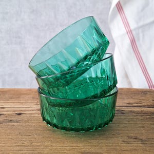 Könnte beinhalten: Ein Stapel aus drei durchscheinenden smaragdgrünen Glasschalen mit einem geometrischen Facettenmuster auf der Außenseite. Die Schalen haben einen kreisförmigen Rand und einen strukturierten Boden, wobei die oberste Schale schräg steht.