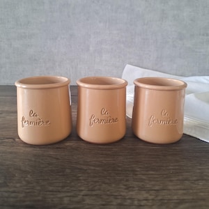 以下が含まれることがあります： 「La Fermière」の文字がエンボス加工された、ベージュ色の陶器製のジャーが3つ。丸みを帯びた形状で、縁がわずかに広がっています。木製の表面に配置されています。