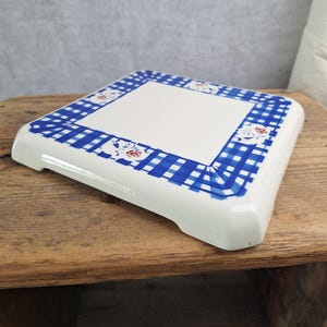 May include: A square ceramic trivet with a white centre and a blue and white chequered border. The border features small, repeating floral motifs in red and white, and the piece has rounded corners and a glossy finish on its top surface.