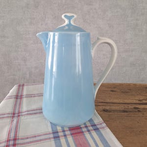 Peut inclure: Une cafetière en céramique bleu clair avec une anse blanche, un bec verseur blanc et un couvercle assorti doté d'un bouton blanc. Le récipient présente une finition lisse et brillante ainsi qu'un corps cylindrique fuselé, avec une anse courbée.