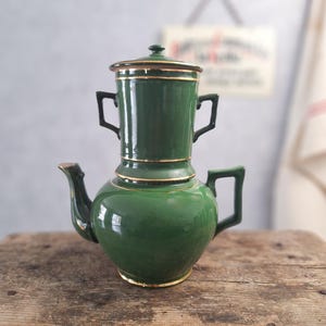 Könnte beinhalten: Eine grüne Vintage-Kaffeekanne aus Keramik mit goldenen Verzierungen. Die Kanne hat einen bauchigen Boden und einen zylindrischen oberen Teil mit zwei Griffen. Ausguss und Griff sind schwarz. Die Kanne steht auf einer verwitterten Holzoberfläche.