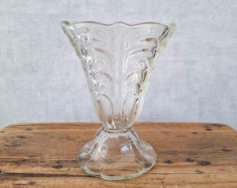 May include: A clear glass vase featuring a flared, scalloped rim and a textured, leaf-like pattern embossed along the exterior body. The vessel stands on a circular, pedestal-style base with a matching scalloped edge design.