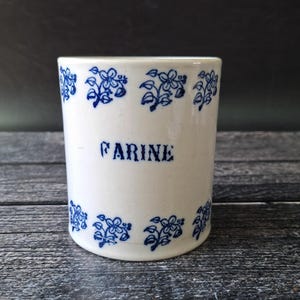 Puede incluir: Un bote de cerámica blanco con patrones florales azules en la parte superior e inferior. La palabra "FARINE" está impresa en azul en la parte delantera. El bote es cilíndrico y está sobre una superficie de madera.