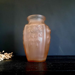 Könnte beinhalten: Eine gefrostete Glasvase in blassrosa mit einem geprägten Design aus Rosen und Blättern. Die Vase hat einen abgerundeten Boden und eine leicht ausgestellte Öffnung.