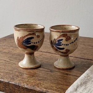 Peut inclure: Deux gobelets en céramique assortis avec un émail crème moucheté. Chaque gobelet est orné de motifs floraux peints à la main dans des tons orange rouille et bleu profond, avec une base en piédestal robuste et un bord arrondi.