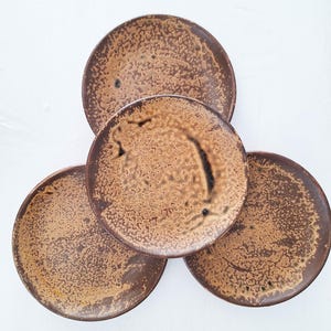 May include: A set of four round ceramic plates arranged in a cluster. Each plate features a textured, mottled glaze in shades of earthy brown and tan, with darker brown speckles and irregular, organic patterns across the surface of the clay.