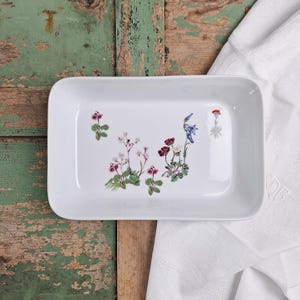Peut inclure: Un plateau de service rectangulaire en céramique blanche orné d'illustrations florales dans les tons de rose, violet et bleu. Le plateau est posé sur une surface en bois rustique avec un linge blanc.