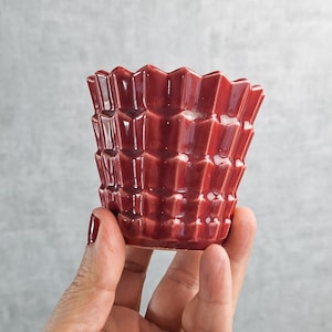 May include: A small, red ceramic planter with a geometric design. The planter has a textured, faceted surface and a slightly flared rim. The color is a deep, rich red, and the planter is held in a hand against a gray background.