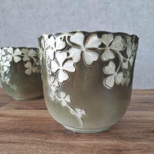 May include: Two ceramic planters with a scalloped rim, featuring a gradient olive green glaze and embossed white clover leaf patterns that wrap around the exterior. The vessels have a rounded, tapered shape and a smooth, glossy finish.