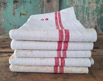 May include: A stack of five folded off-white linen napkins featuring two vertical red stripes and the embroidered red monogram BC on the top piece. The fabric has a visible woven texture with light tan discoloration spots throughout.