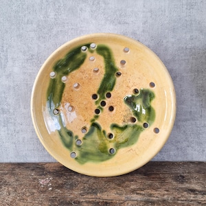 Puede incluir: Un jabonera de cerámica hecha a mano con forma redonda. El plato presenta una base amarilla con un diseño verde de forma orgánica y múltiples orificios de drenaje. El plato está colocado sobre una superficie de madera.