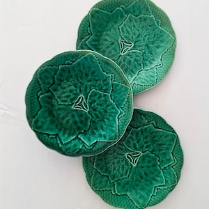 May include: Three green ceramic plates with a leaf design. The plates have a textured surface and a scalloped edge. The plates are arranged on a white surface. The plates are a deep shade of green.