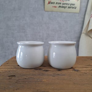 Może przedstawiać: Dwa małe, białe ceramiczne słoiczki o zaokrąglonych korpusach i lekko rozszerzających się krawędziach stoją na drewnianej powierzchni. Słoiczki są gładkie i błyszczące, odbijając światło. W tle wisi szyld z francuskim tekstem.