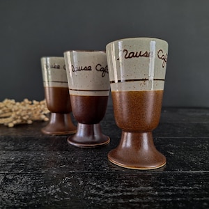 May include: Three brown and white ceramic coffee cups with a brown base and the words "Pause Cafe" written in brown on the white part of the cup.