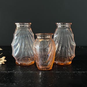 May include: Three vintage pink glass vases. Two vases have a fluted design, and the third is a smaller, rounded vase with a spiral pattern. The vases are arranged on a dark surface, with a black background.
