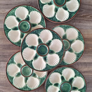 Könnte beinhalten: Sechs Austernplatten aus Keramik. Jede Platte hat ein grün-weißes Muscheldesign mit einer zentralen Vertiefung. Die Platten haben einen strukturierten, gewebten Rand und einen braunen Rand. Die Platten sind auf einer Holzoberfläche angeordnet.