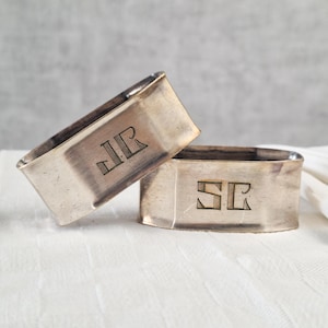 May include: Two silver napkin rings with engraved initials. One ring displays "JR" and the other "SR". The napkin rings are rectangular with slightly rounded edges. They are placed on a white, textured surface.