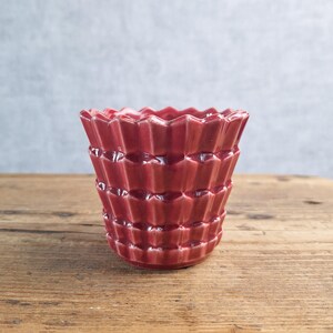 May include: A deep red ceramic planter featuring a distinct geometric, pleated, or accordion-like texture across its exterior surface. The vessel has a wide, scalloped rim and a tapered body, showcasing a glossy finish that reflects light.
