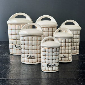 Peut inclure: Ensemble de six pots en céramique blanche avec des garnitures dorées et des étiquettes. Les étiquettes indiquent "Suc", "Epices", "Poivre", "The" et "fe".